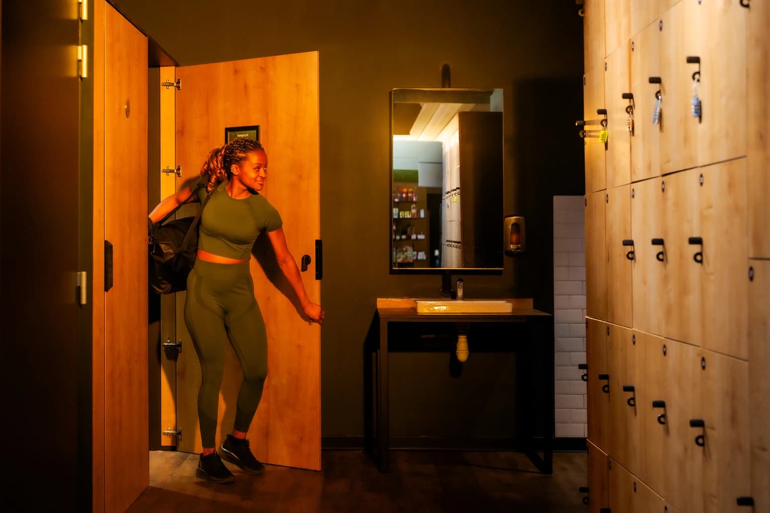 Vestiaire salle de sport avec femme en tenue d’entraînement devant rangements et miroir.